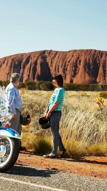 Three travellers park their motorcycle at the side of the road, in view of Ayers rock